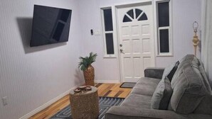 Un salón con un sofá gris, un televisor de pantalla plana montado en la pared, una puerta blanca con paneles de vidrio y una pequeña mesa redonda con una planta.
