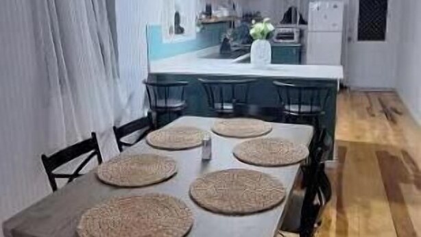 Un comedor con una mesa y sillas de madera, una cocina al fondo y una cortina blanca a la izquierda.