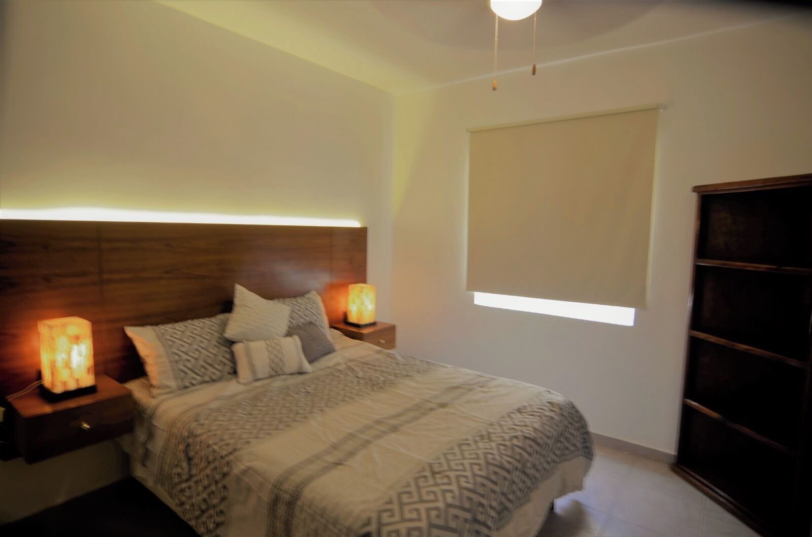 Une chambre à coucher avec une tête de lit en bois, deux lampes de chevet, un lit avec une literie à motifs, une table de chevet, une armoire et une fenêtre avec des stores.