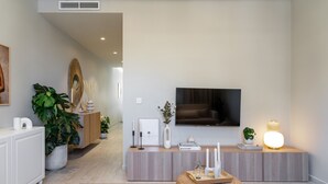 Un salon moderne avec un téléviseur à écran plat fixé au mur, une console en bois agrémentée d’objets décoratifs, un pouf en osier et une grande plante.