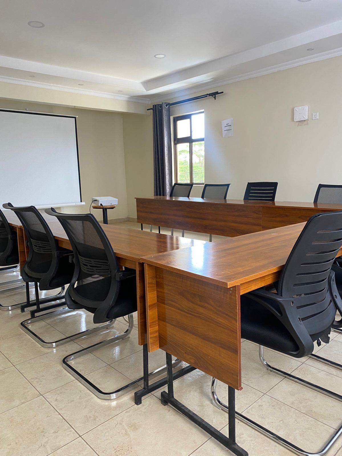 Une salle de conférence avec une longue table en bois, des chaises noires à dossier en résille, un écran de projection et une fenêtre avec des rideaux.