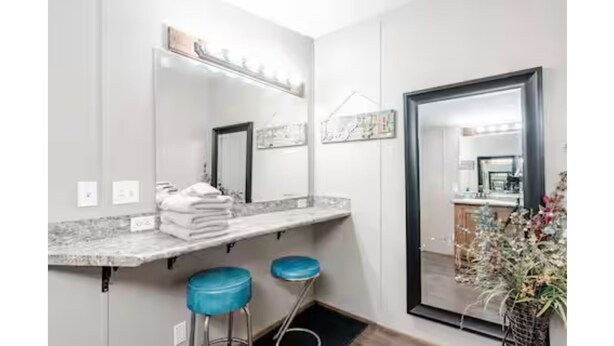 A modern bathroom with a marble countertop, two blue bar stools, a large mirror, and a potted plant.