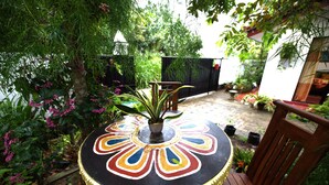 Taula rodona amb una superfície pintada i colorida, envoltada per una cadira de fusta, a un jardí amb diverses plantes i una tanca al fons.