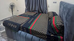 A bedroom with a bed featuring patterned bedding and pillows, a standing fan, and a ceiling light. - Smart 2-bedroom bungalow in lovely Egbeda with WiFi, AC,parking space,games etc… (Egbeda)