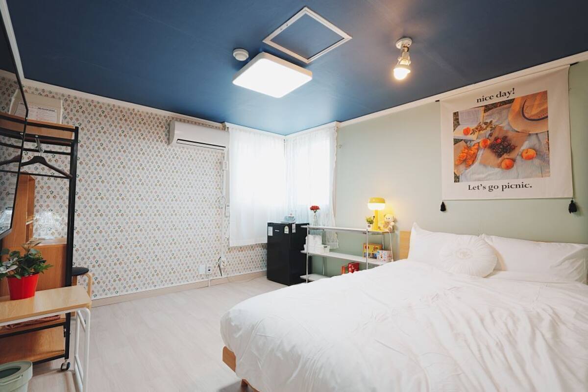 A bedroom with a bed, a desk with a plant, a radiator, a window with curtains, and a poster on the wall.