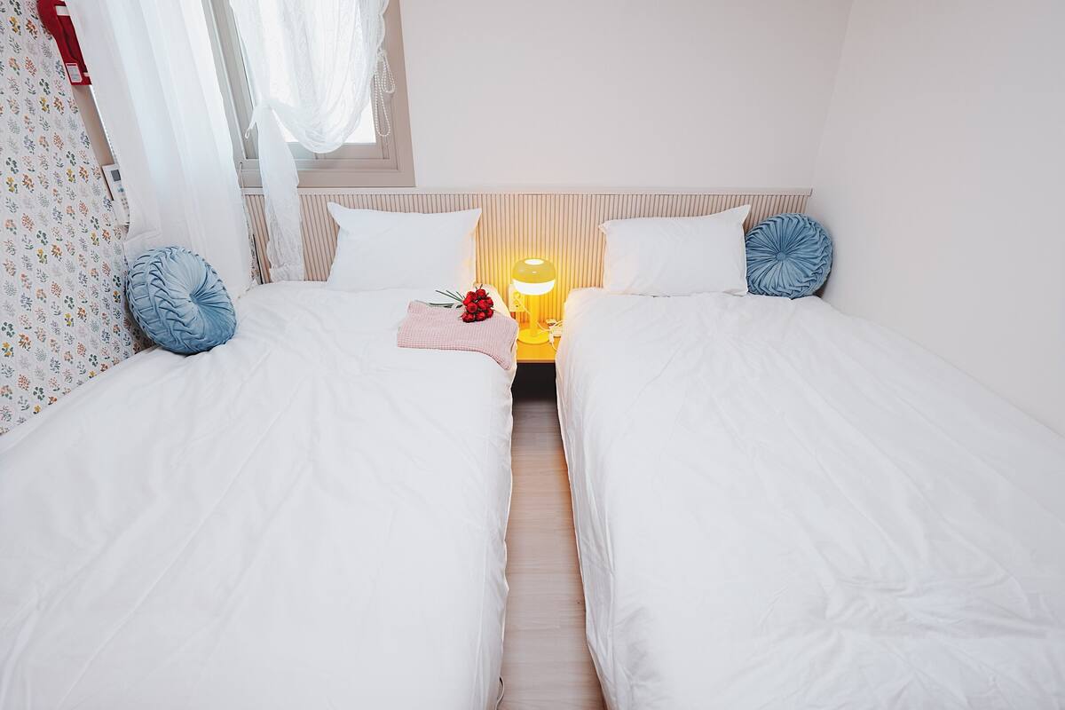 A hotel room with two single beds, a bedside table with a lamp, and a window with sheer curtains.