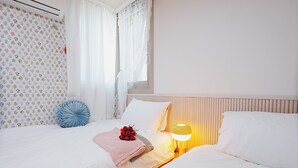 Een netjes opgemaakt bed met witte lakens, een blauw kussen, een roze deken en een boeket rode rozen. Er staat een bedlamp met een gele lampenkap en een raam met dunne gordijnen.