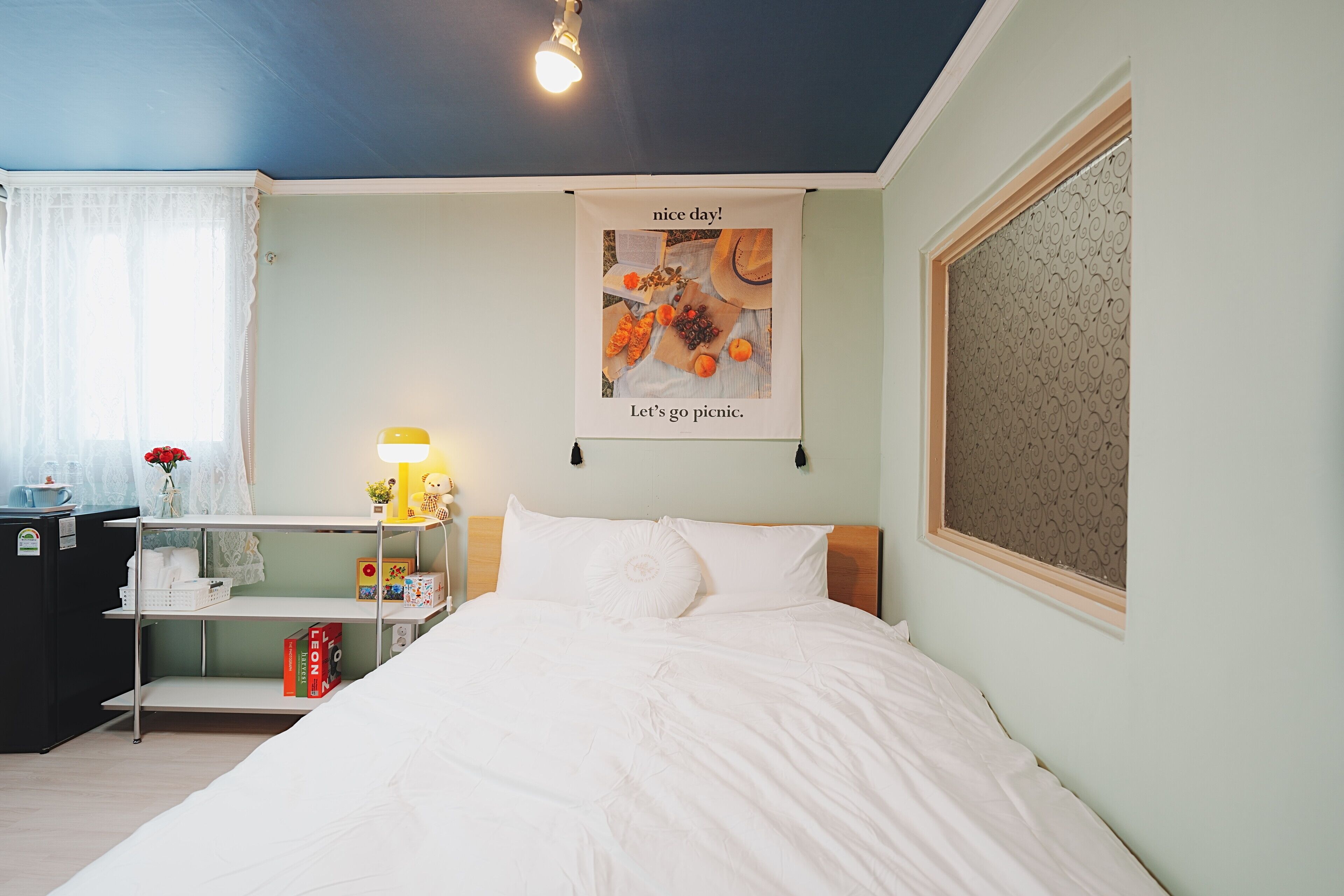 A bedroom with a bed, a bedside table with a lamp, a poster on the wall, and a window with a curtain.