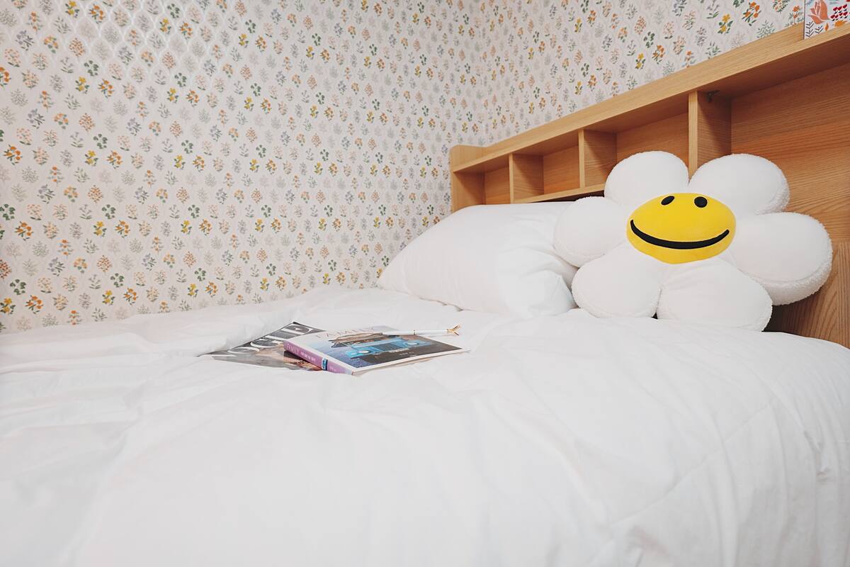 A bed with a white floral-patterned wall, a wooden headboard, and a large flower-shaped pillow with a smiley face.