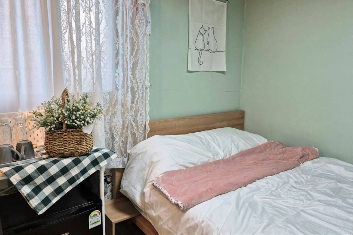 A bedroom with a bed, a nightstand, a lamp, a basket with flowers, and a wall art of two cats.