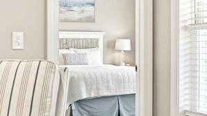 Een slaapkamer met een bed, een bedlamp, een kussen met de tekst 'BEACH' en een schilderij aan de muur.
