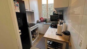 A compact kitchen with white cabinets, a black refrigerator, a microwave, and a small window. - Cosy near the Eiffel Tower (Paris)
