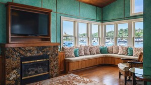 Een kamer met een ingebouwde televisie, een stenen open haard, een gebogen zithoek met gekleurde kussens en grote ramen met uitzicht op een jachthaven.