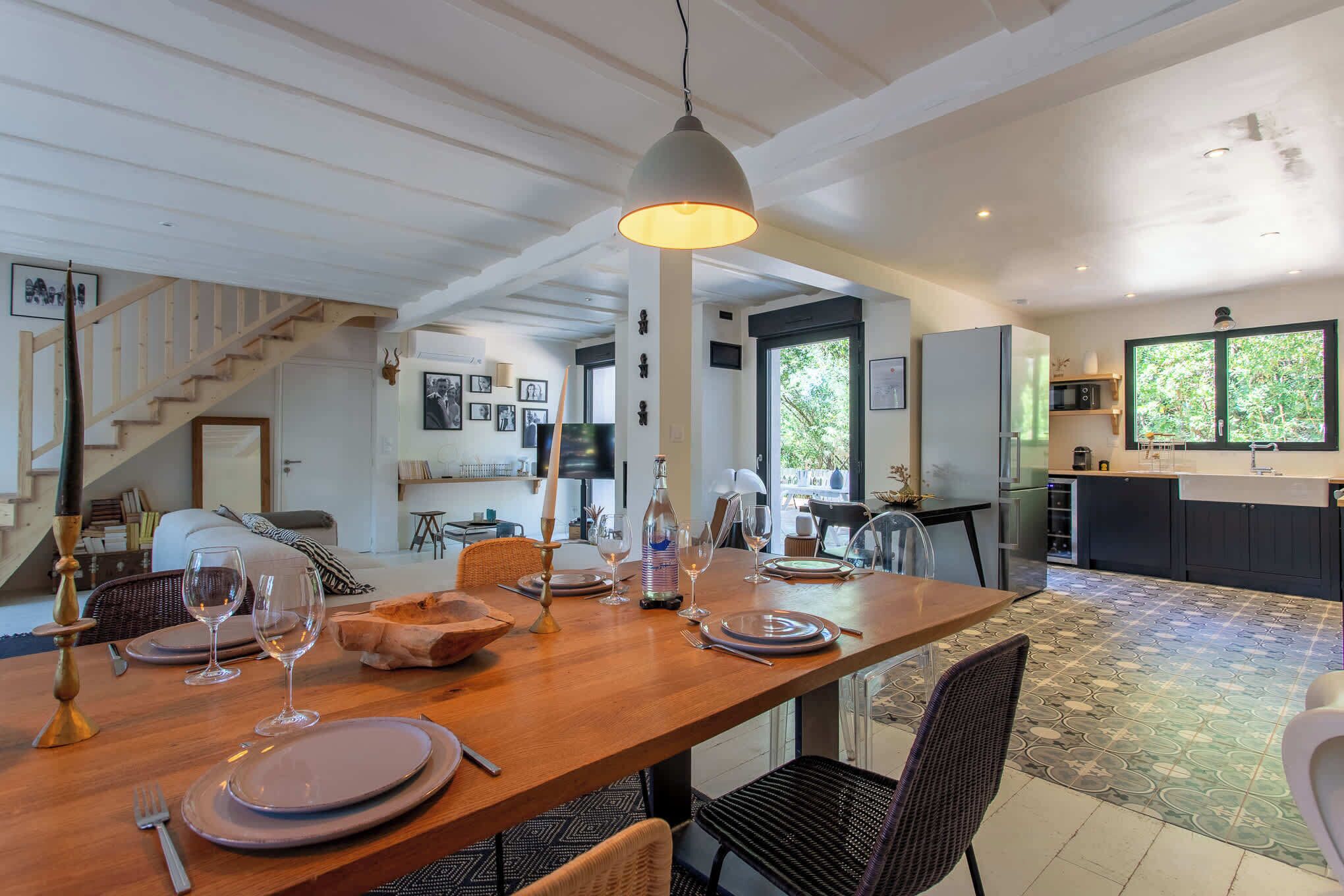 Uma sala de jantar com mesa de madeira posta para quatro pessoas, luminária pendente e vista para a cozinha e sala de estar ao fundo.