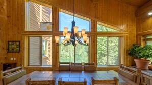 A wooden interior with a large dining table and benches, a chandelier, and large windows offering a view of the outdoors.