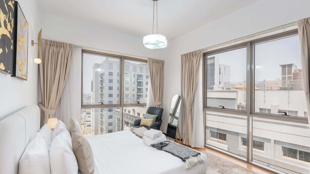 A modern bedroom with a large bed, a chair, and a view of the cityscape through large windows. - StayShort - Spacious 2-Bed Al Barsha Escape - Balcony & Modern Amenities, Fits 4 (Al Barsha 1)