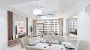 A modern dining area with a large table set for four, a television on the wall, and a view of the cityscape through large windows. - StayShort - Spacious 2-Bed Al Barsha Escape - Balcony & Modern Amenities, Fits 4 (Al Barsha 1)