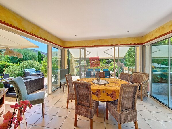 Une terrasse ensoleillée avec une table à manger, des chaises en osier et une porte vitrée donnant sur un jardin.
