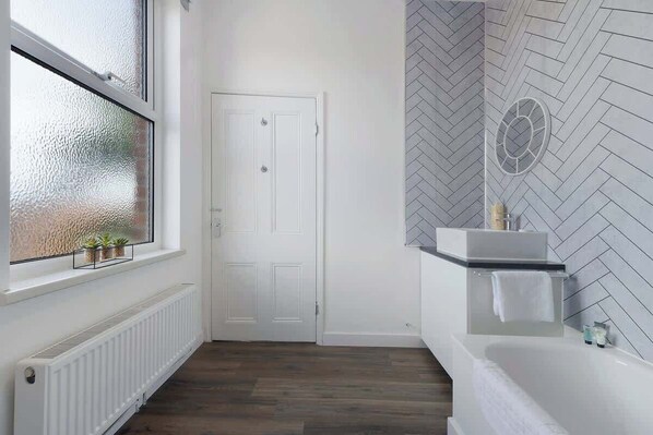 A bathroom with a herringbone-patterned wall, a white door, a window with frosted glass, and a white bathtub.
