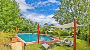 Piscina retangular com deck ao redor, cadeiras de sol e uma pérgula que oferece sombra.