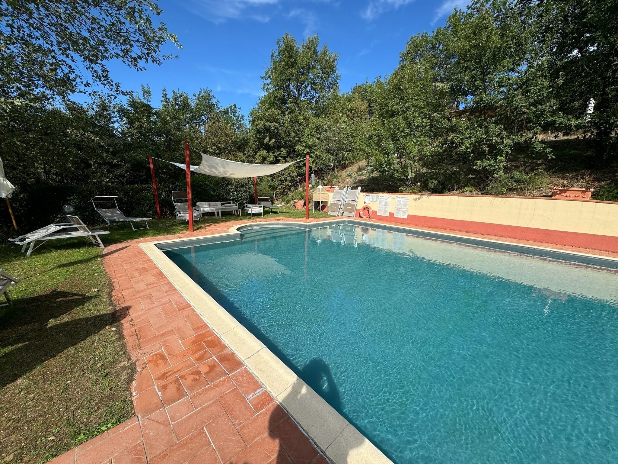 Une piscine rectangulaire dont les bords sont recouverts de carreaux, entourée d’une terrasse en briques. On y trouve des chaises longues et un espace ombragé avec un auvent.