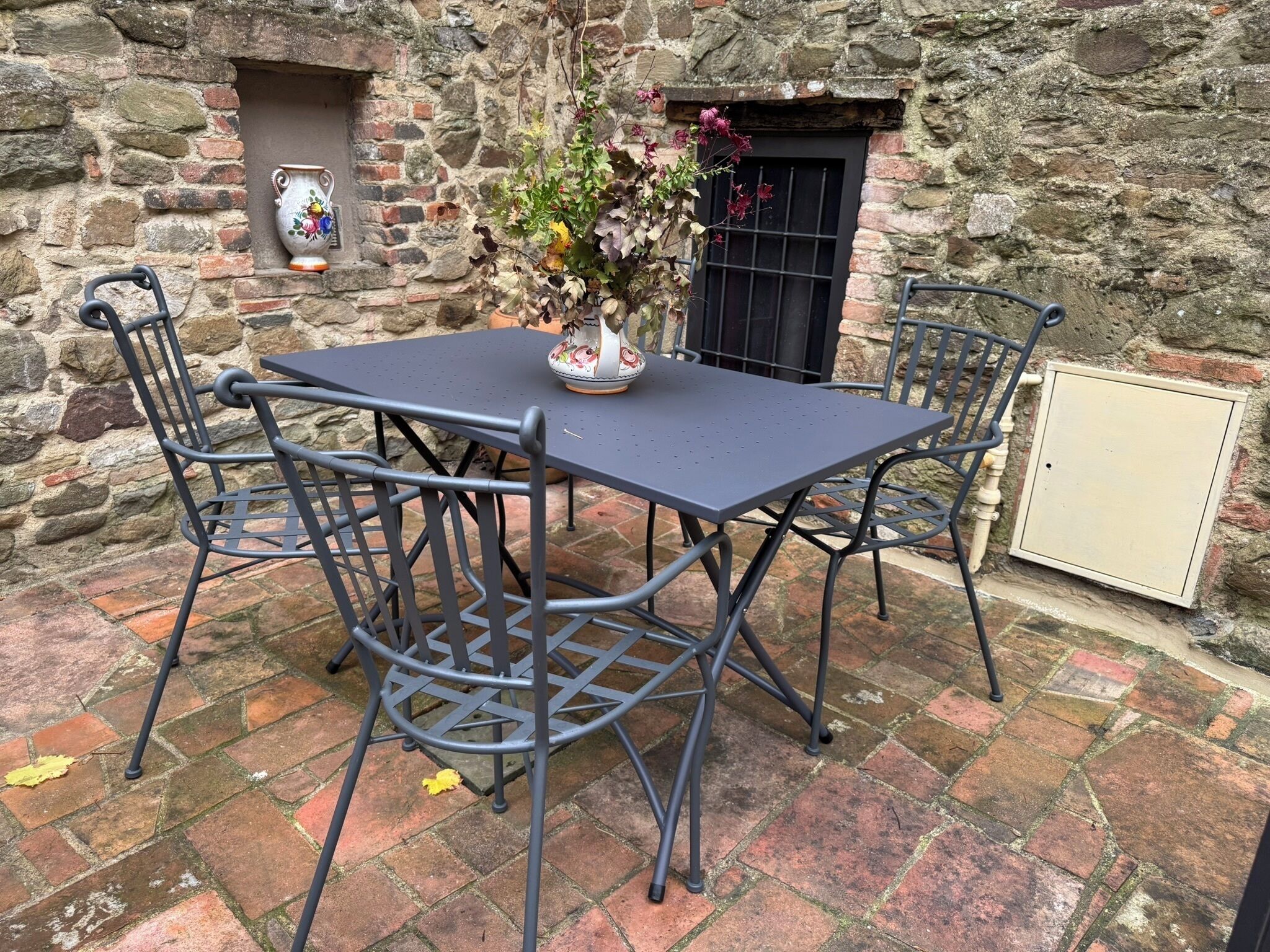 Un mur de pierre avec une table et des chaises en métal, un vase de fleurs et une petite fenêtre.