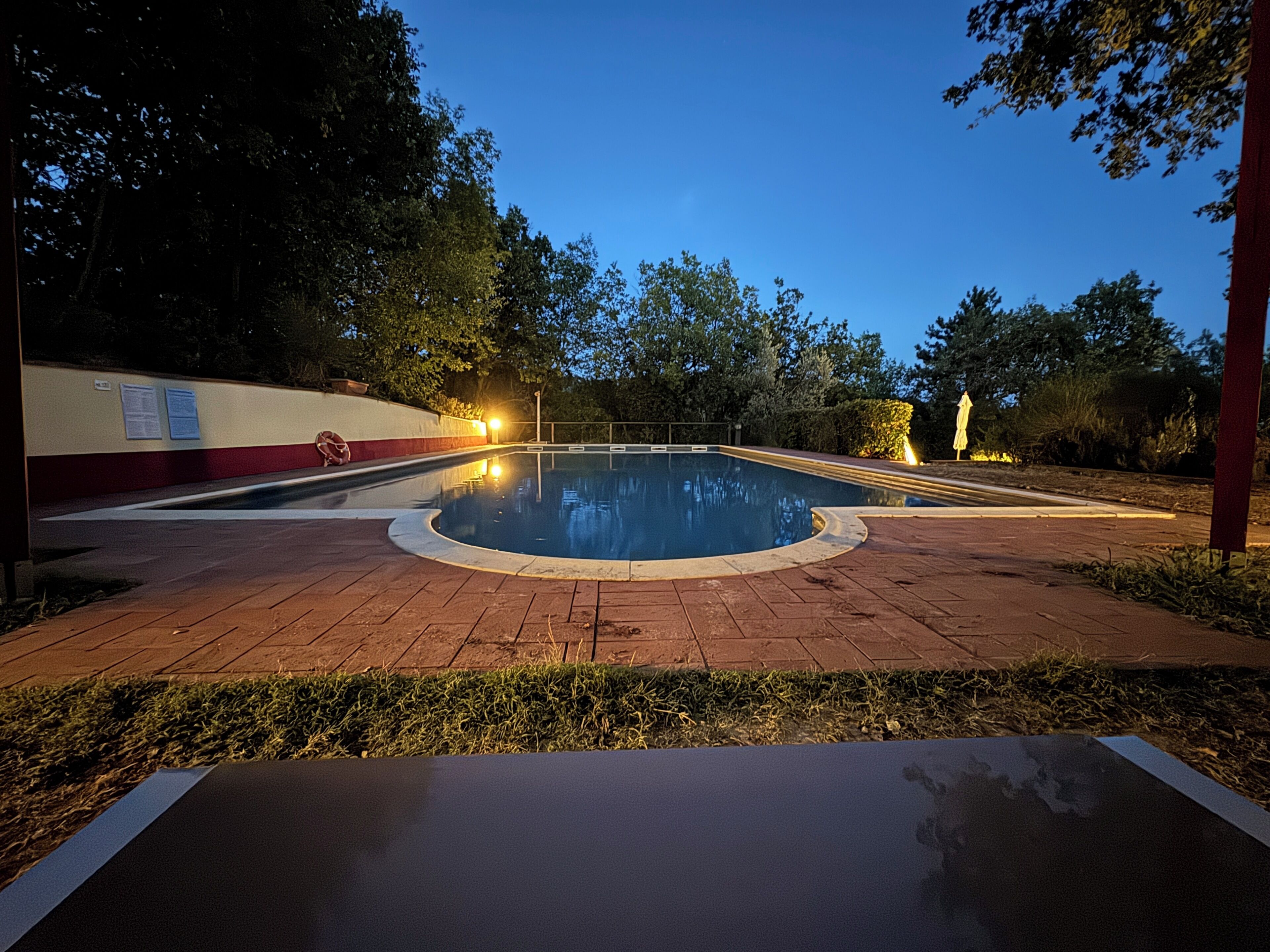 A swimming pool with a paved deck, surrounded by trees and a red wall with informational signs.