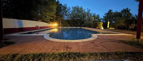 A swimming pool with a paved deck, surrounded by trees and a red wall with informational signs.