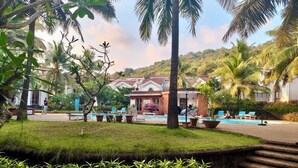 A resort with a swimming pool, palm trees, and a building with a red roof. - Goan Hideaway - Tres (Arpora)