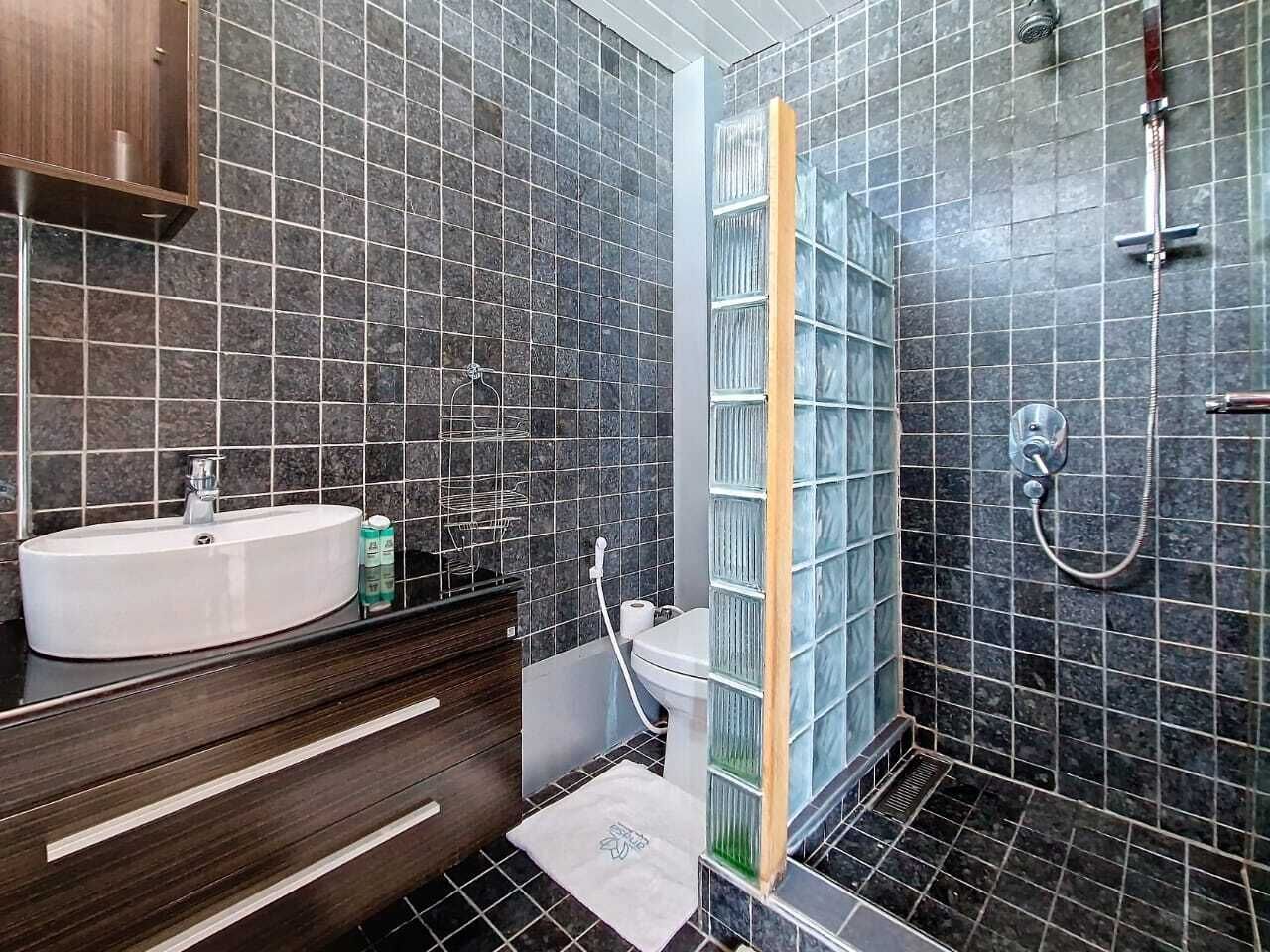A modern bathroom with a white sink, dark tiled walls, a glass shower enclosure, and a wooden vanity.