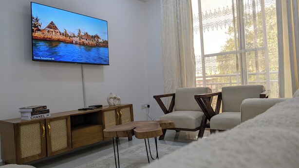 A living room with a TV displaying a scenic relaxation program, a wooden cabinet with books, a small table, and two chairs. - Kays Place Zanzibar (ZANZIBAR)