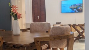 A dining area with a wooden table and chairs, a vase with flowers, a television on the wall, and a ceiling fan. - Kays Place Zanzibar (ZANZIBAR)