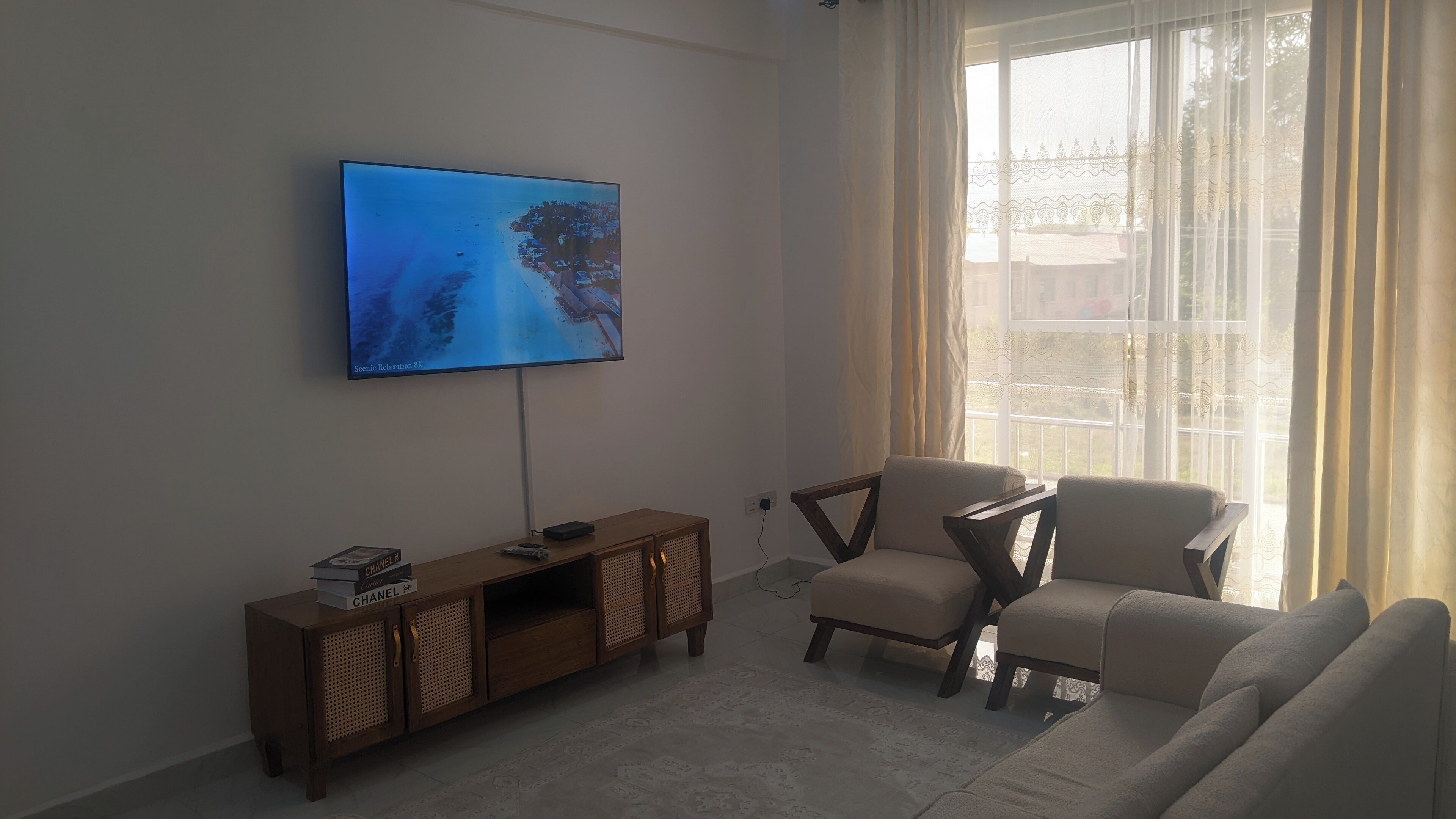 A living room with a flat-screen TV mounted on the wall, a wooden TV stand with books, a beige sofa, and a window with sheer curtains.