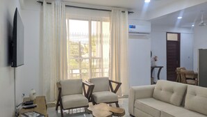 A living room with a sofa, two armchairs, a wooden coffee table, a TV mounted on the wall, and an air conditioning unit. - Kays Place Zanzibar (ZANZIBAR)