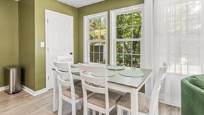 A dining area with a white table and chairs, a green wall, and a window with a view of greenery.