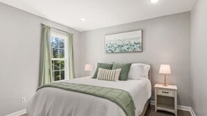 A bedroom with a bed, two bedside tables, a window with curtains, and a wall-mounted artwork.