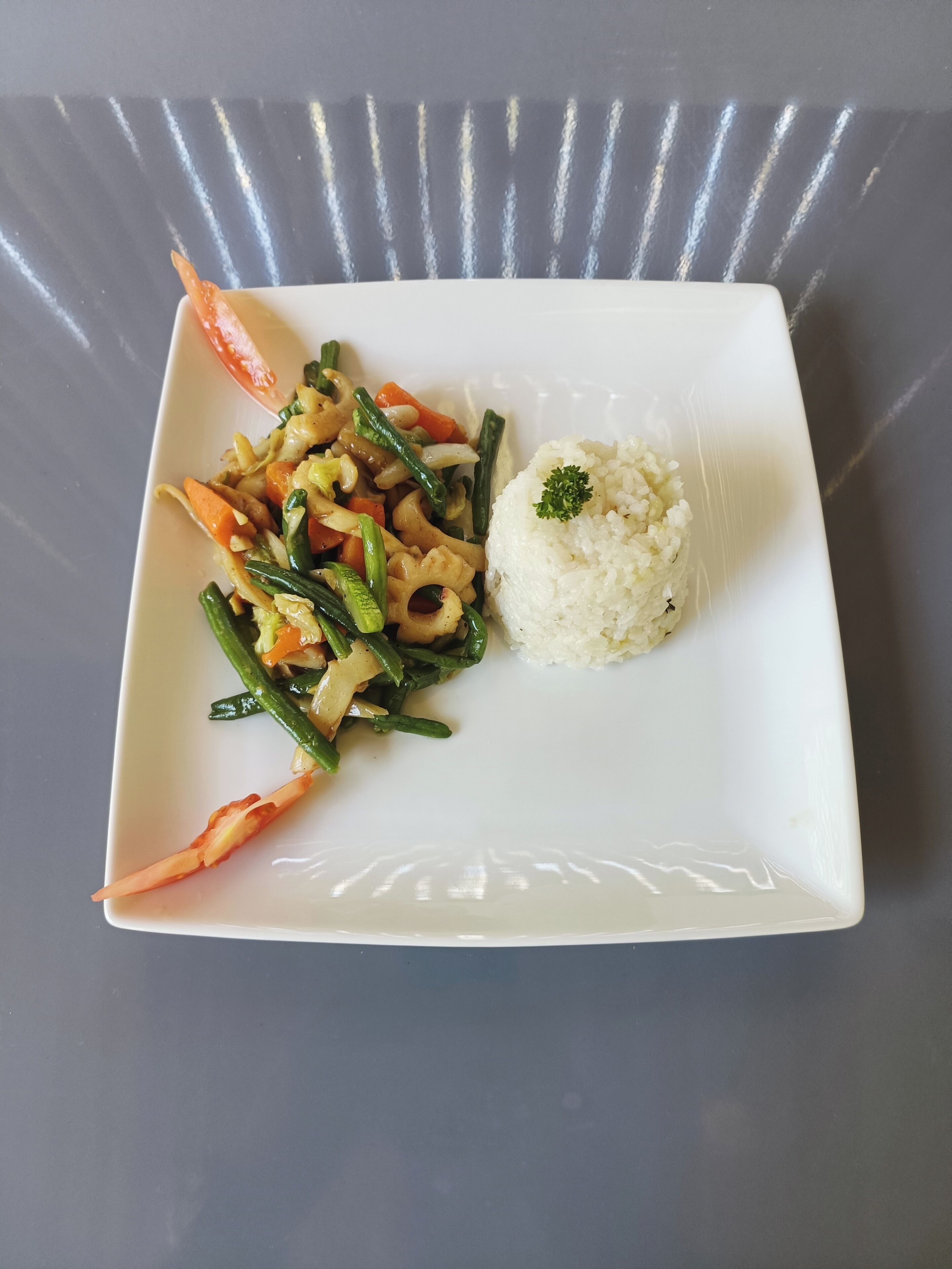 A square plate with a stir-fry dish and a mound of white rice, garnished with a green leaf.