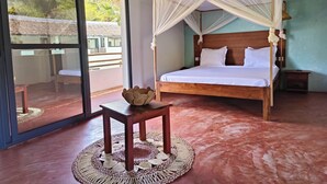 A room with a bed, a small table, a woven basket, and a view through a glass door. - SAKATIA ART (Nosy Be)