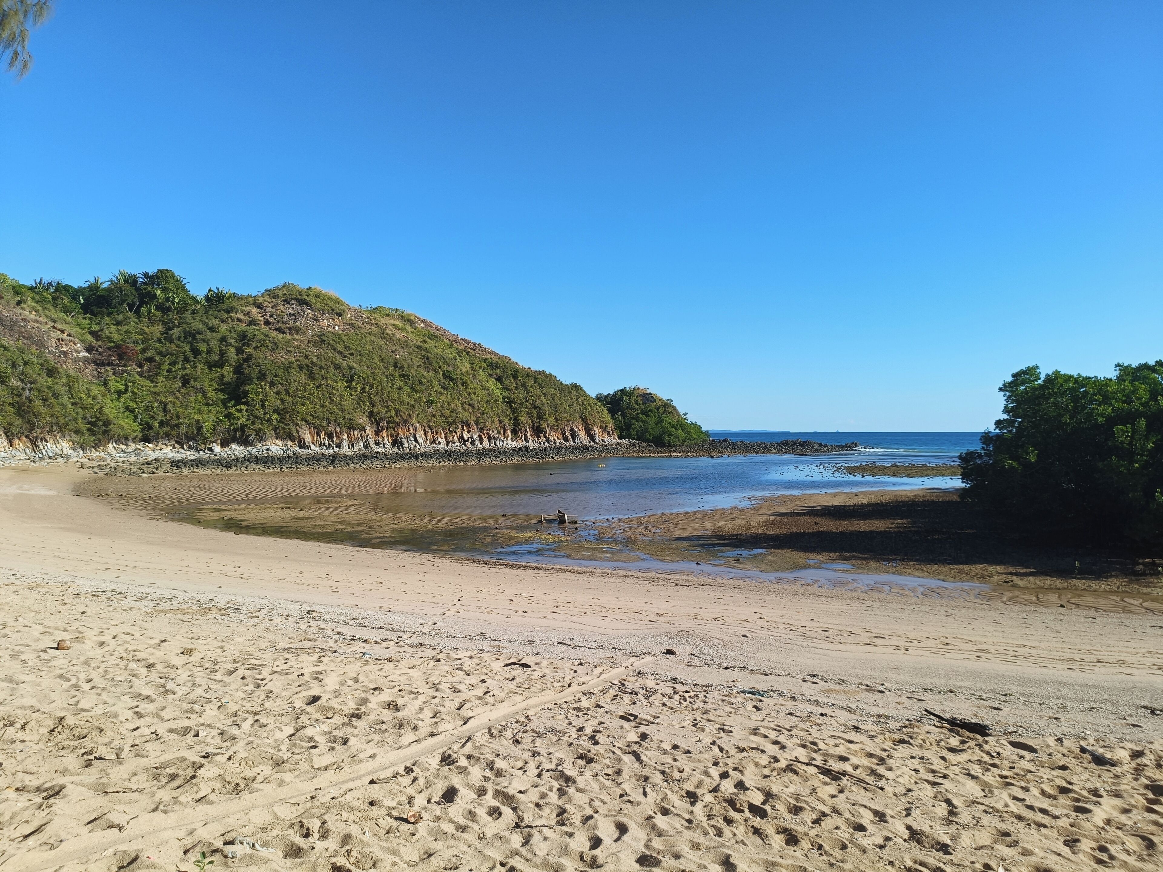 Ein Strand mit felsiger Küste, blauem Himmel und einer bewaldeten Hügelkulisse im Hintergrund.