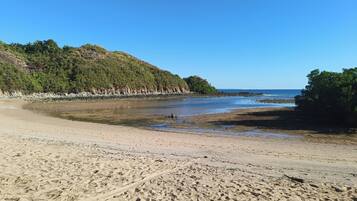 Ein Strand mit felsiger Küste, blauem Himmel und einer bewaldeten Hügelkulisse im Hintergrund.