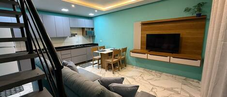 Una sala de estar moderna con un sofá, una mesa de comedor con sillas, una zona de cocina y un televisor fijado a la pared.