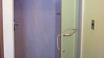 Ducha con mampara de vidrio, cabezal de ducha de mano, puerta de vidrio con manija de metal y un estante pequeño con artículos de aseo.