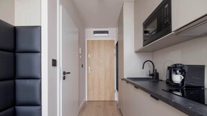 Un pasillo estrecho que conduce a una puerta de madera, con un banco acolchado negro a la izquierda y una cocina a la derecha que incluye un microondas, una cafetera y una encimera.