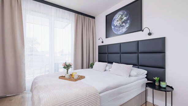 Un dormitorio moderno con una cama grande, cabecera, mesita de noche, un cuadro en la pared y una ventana con cortinas.