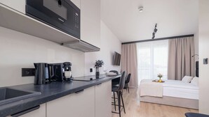 Una cocina moderna con cafetera integrada, un comedor con una mesa y sillas pequeñas, y un dormitorio con cama y cortinas.