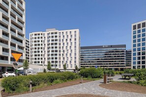 A modern urban area with a multi-story building featuring a UBS sign, surrounded by greenery and a paved walkway.