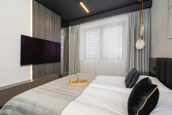 Una habitación de hotel moderna con una cama grande, un televisor de pantalla plana montado en la pared y una mesita de noche con una bandeja que contiene tazas y una pequeña planta.