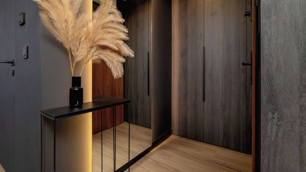 Un pasillo moderno con piso de madera, una consola negra y un florero con plumas de pampa, y puertas de madera oscura.