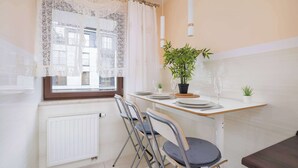 Eine moderne Küche mit Essbereich, weißen Fliesen, einem Fenster mit Spitzenvorhängen, einem Heizkörper und einer Topfpflanze.