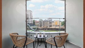 A balcony with two wicker chairs and a small table, overlooking a cityscape with buildings and a playground.